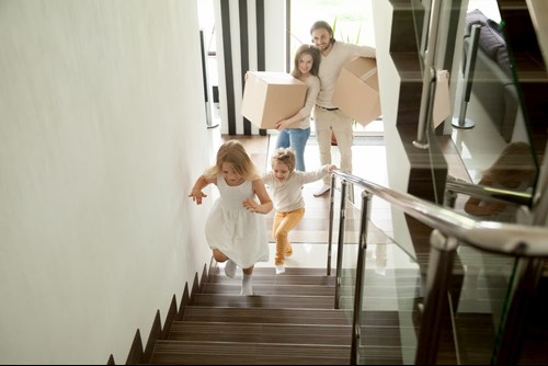 Two children running upstairs to their new home as the parents stand behind them holding moving boxes.